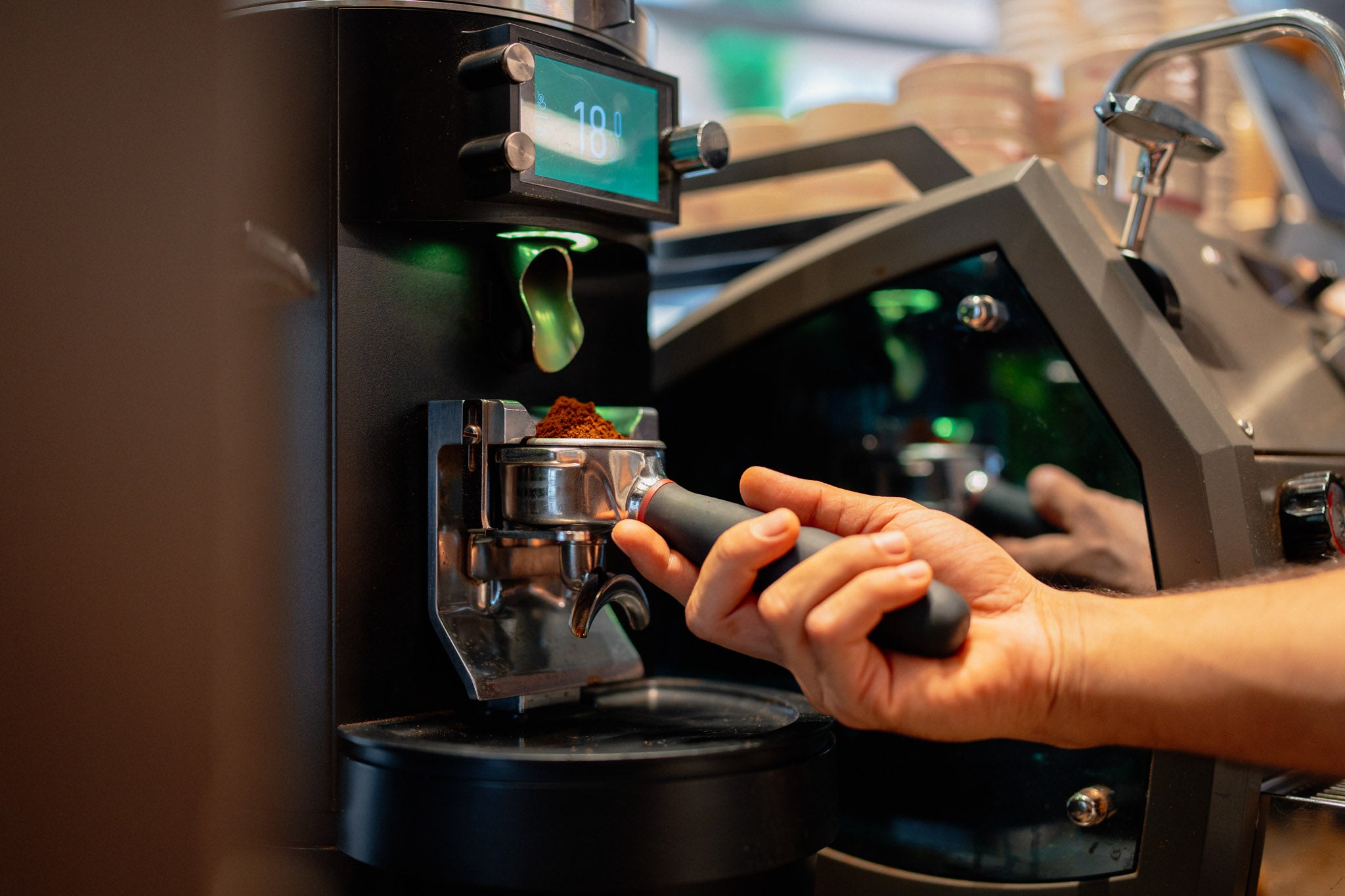 A close-up of a person's hand holding a portafilter under a coffee grinder dispensing freshly ground coffee, with a warm, blurred cafe background.